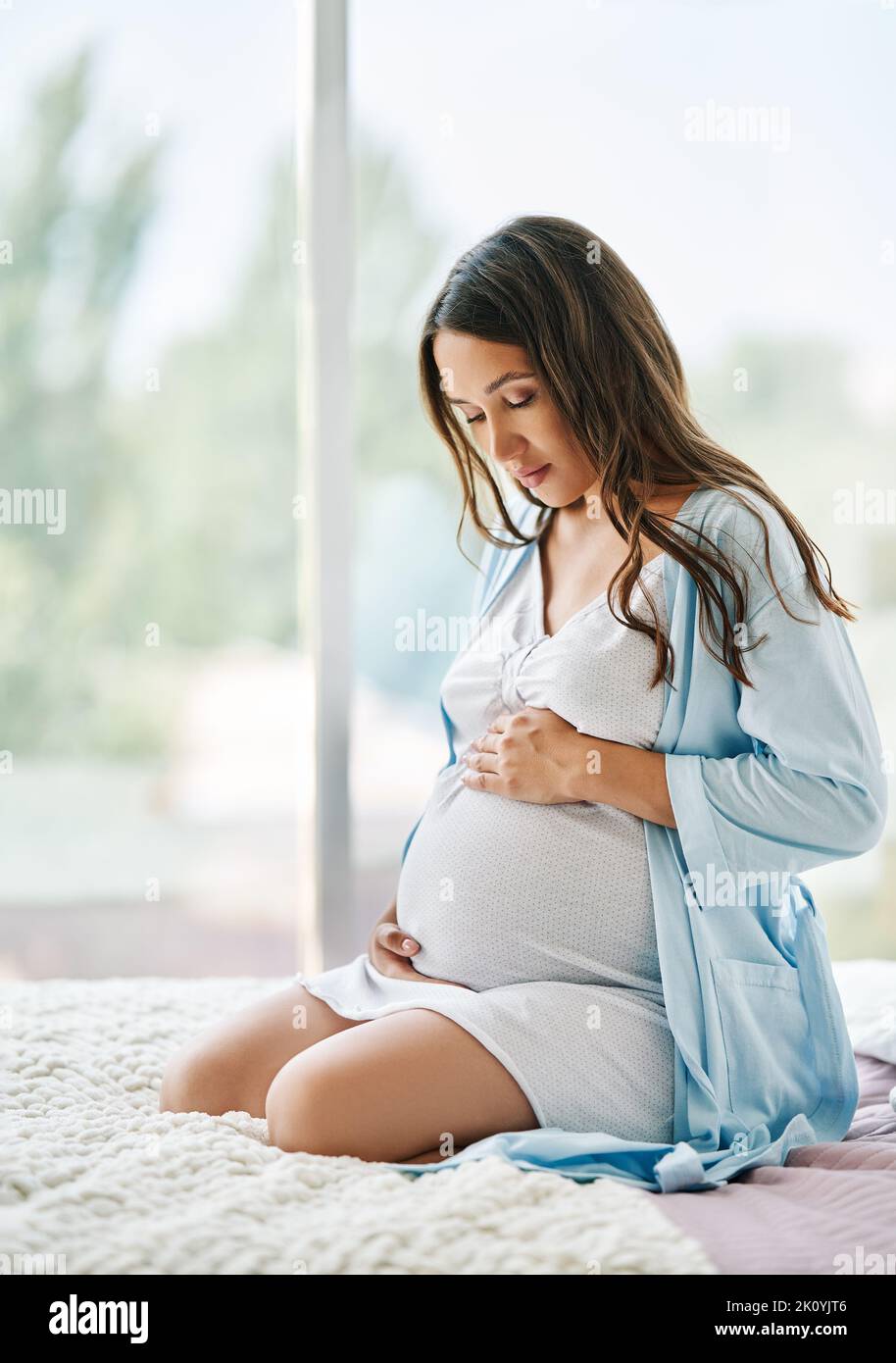 Portrait of young pregnant woman relax sittong in bed and touching her belly at home. Motherhood, people concept. Last months of pregnancy. Stock Photo