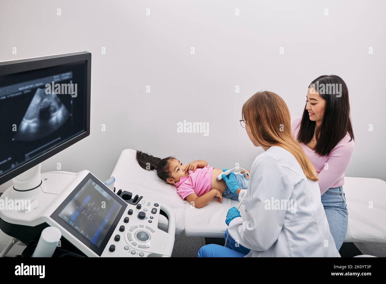 Doctor with ultrasound probe scanning abdominal and internal organs of Asian child using ultrasound machine. Little girl with support of her mother, r Stock Photo