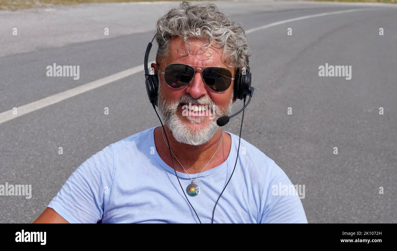 Gray-haired elderly man freelancer with a beard in sunglasses, working in the headphones on road. A crazy and extraordinary old senior in the summer Stock Photo
