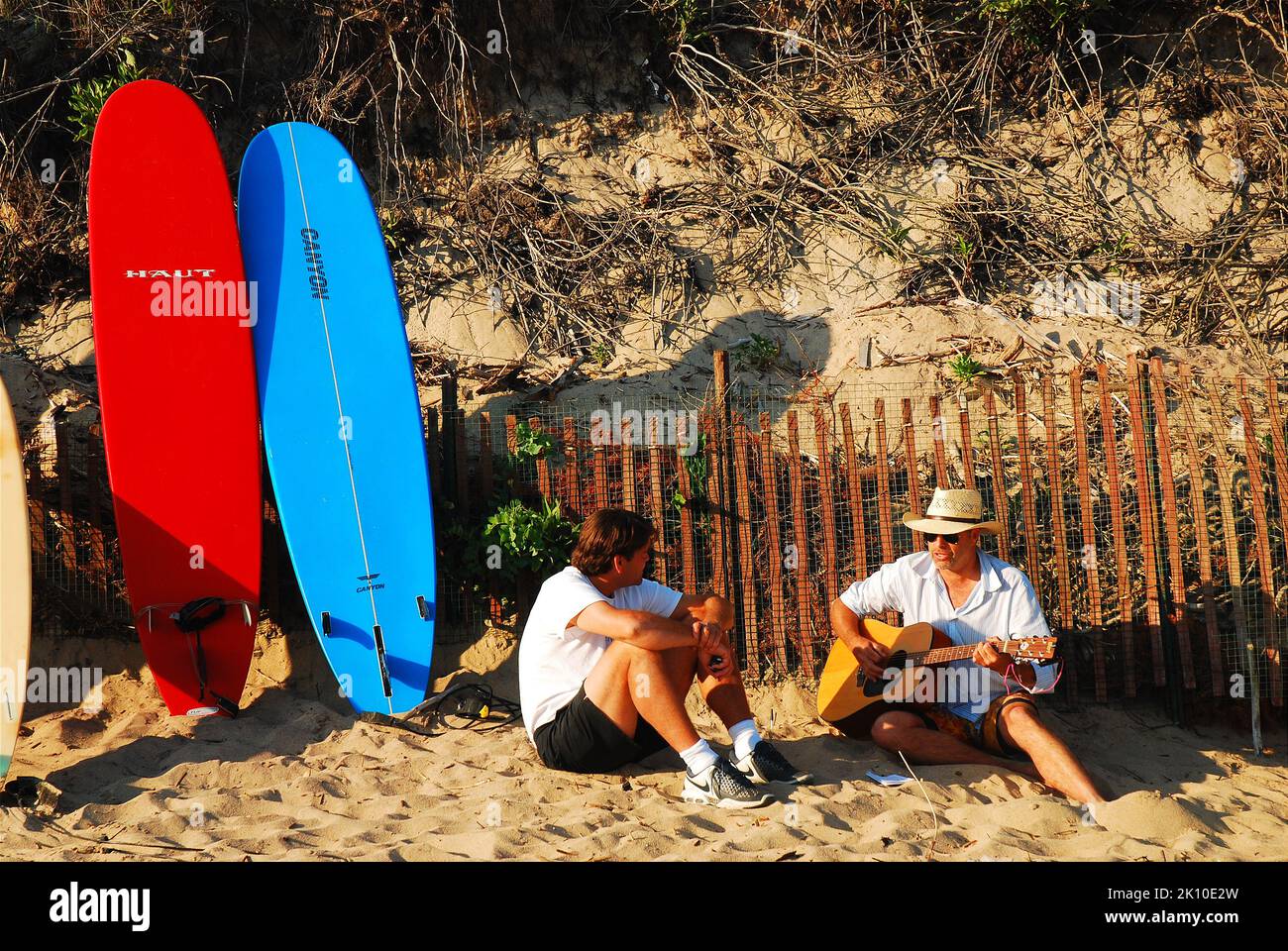 Two adult men sing and play guitar on the beach with their surfboards behinds them on a sunny summer vacation day Stock Photo