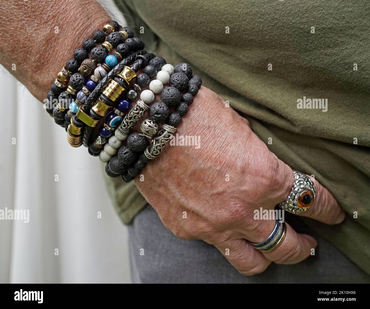 Senior man putting his hand in the pocket of his grey jeans. He wears lava stone and other breaded bracelets and special rings. Stock Photo