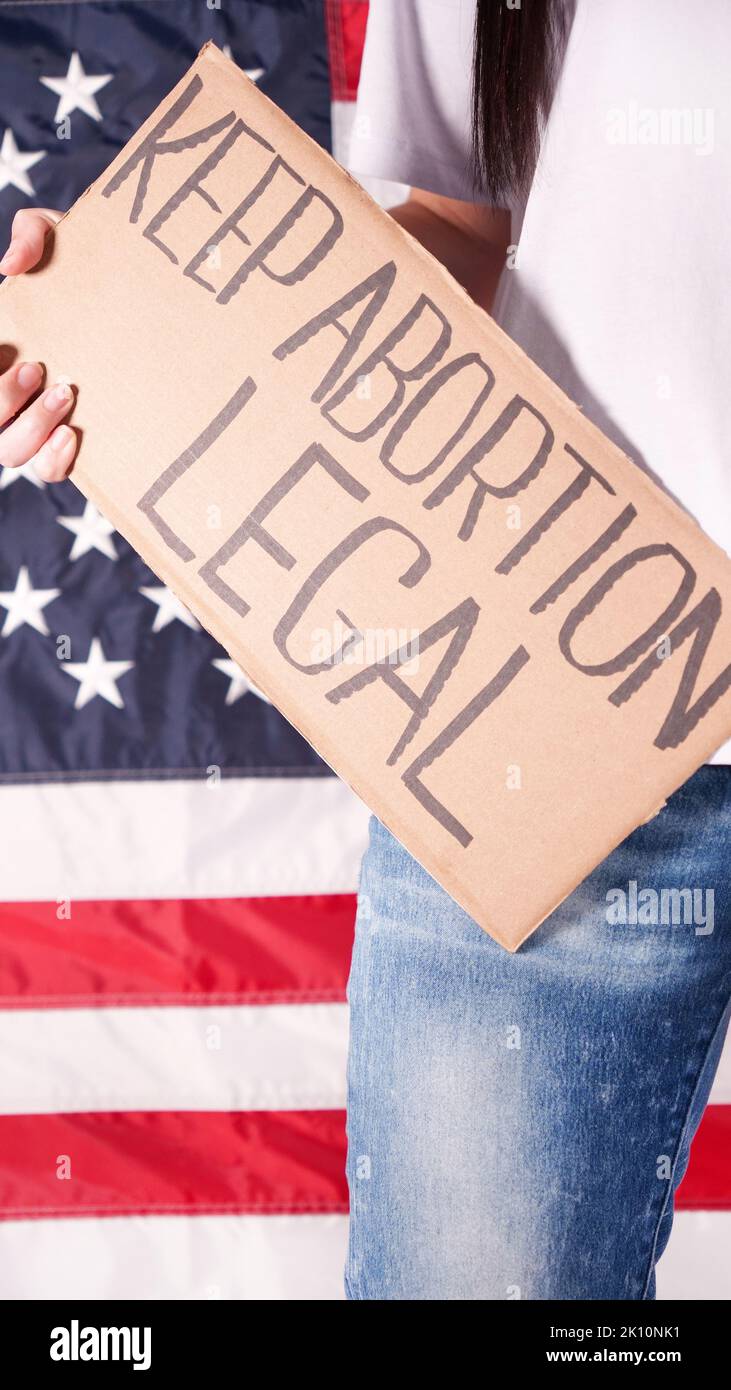 Young woman protester holds cardboard with Keep Abortion Legal sign against USA flag on background. Girl protesting against anti-abortion laws. Femini Stock Photo