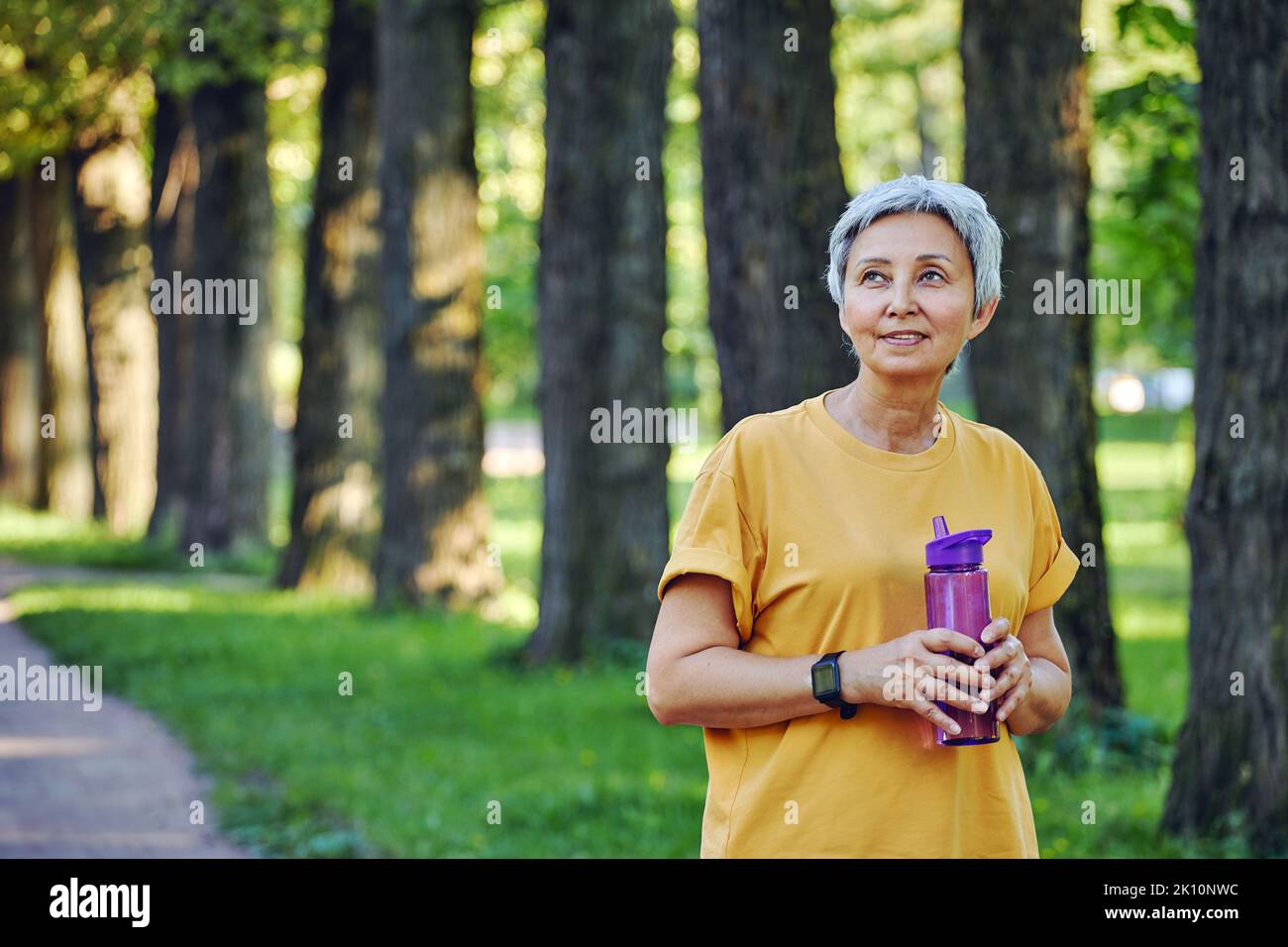 Older woman holds reusable plastic bottle standing in footpath in park summer forest, caring about health, relieving thirst during morning work out. H Stock Photo