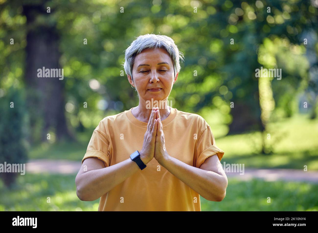 Older active Asian woman do meditation practice with eyes closed pose in summer park folded arms makes mudra or Namaste sign, do yoga work out outdoor Stock Photo