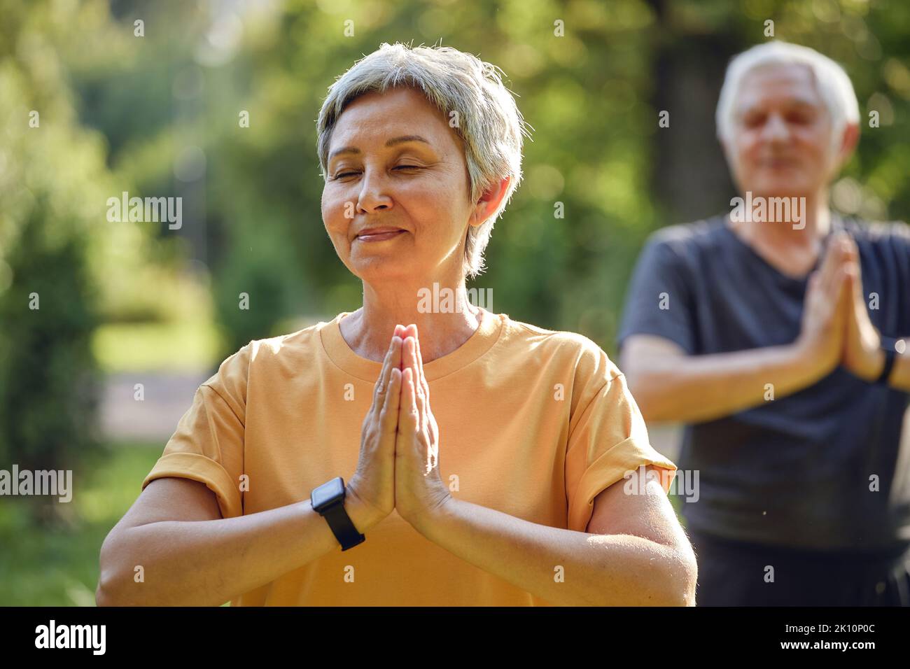 Older active couple do meditation practice with eyes closed pose in summer park folded arms makes mudra or Namaste sign, do yoga work out outdoors, he Stock Photo