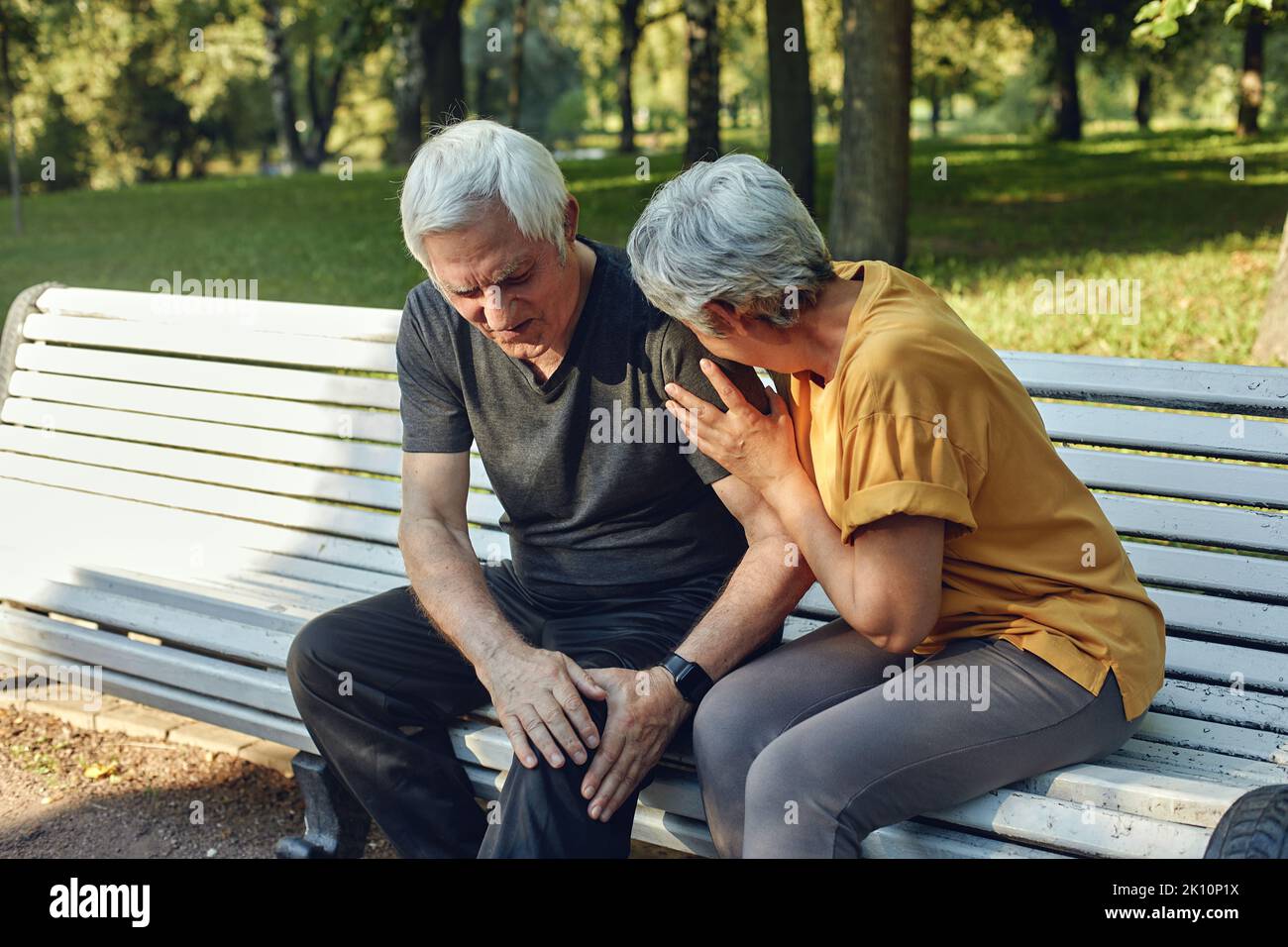 During morning sportive stroll in summer park, elderly 70s man got injured his knee, gripping leg seated in bench with caring disappointed wife. Physi Stock Photo