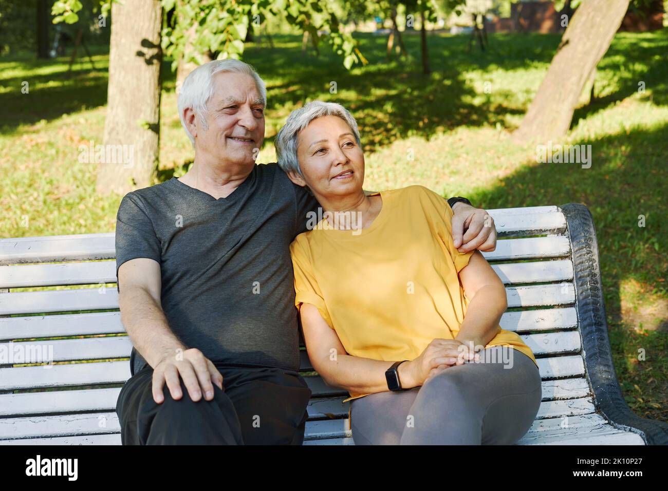 Happy multi racial elderly grey hair couple resting sit on bench in park after stroll, morning walk smiling enjoy relaxation outdoors at sunny summer Stock Photo