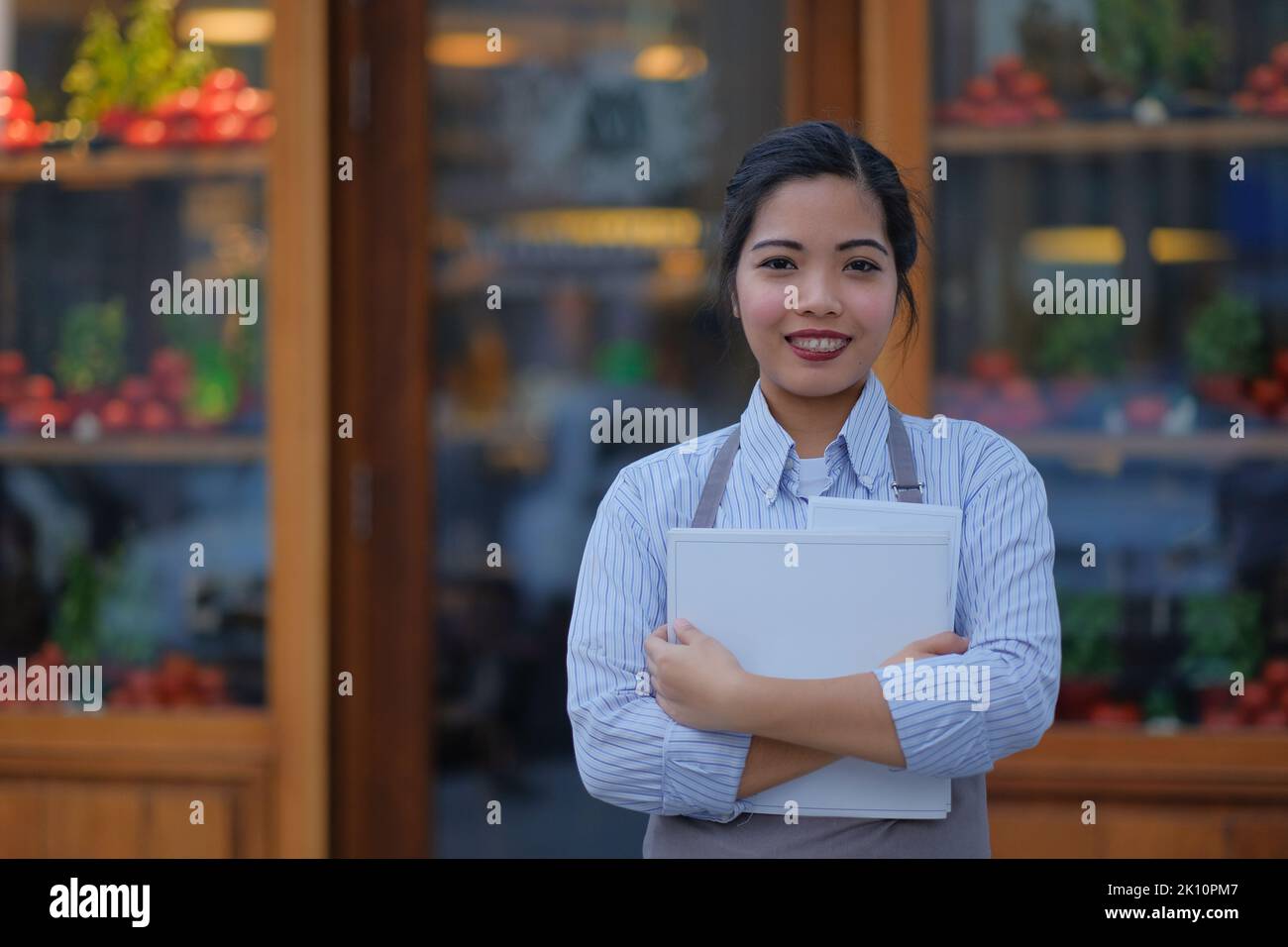 Young Filipino waitress waits outside an Italian restaurant window with menus and a smile. Outdoor portrait of an Asian female in the service industry. Stock Photo