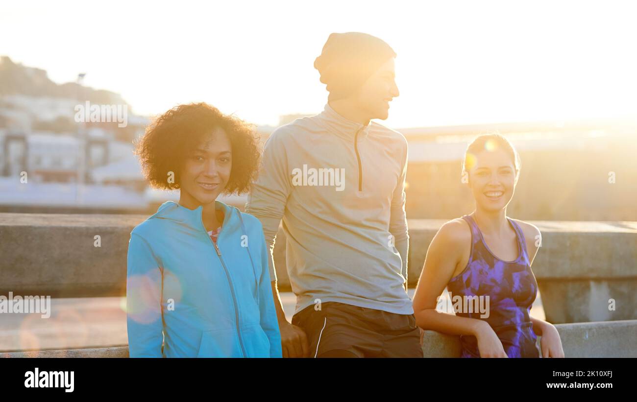 Enjoying an invigorating morning run. Portrait of three friends taking a break at the road side during a morning jog. Stock Photo