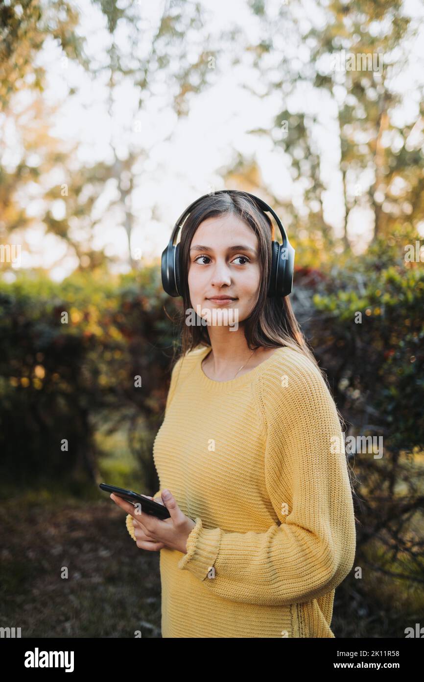 Teenage girl wearing yellow sweater, using headphones for playing music on her smartphone outdoor. Stock Photo