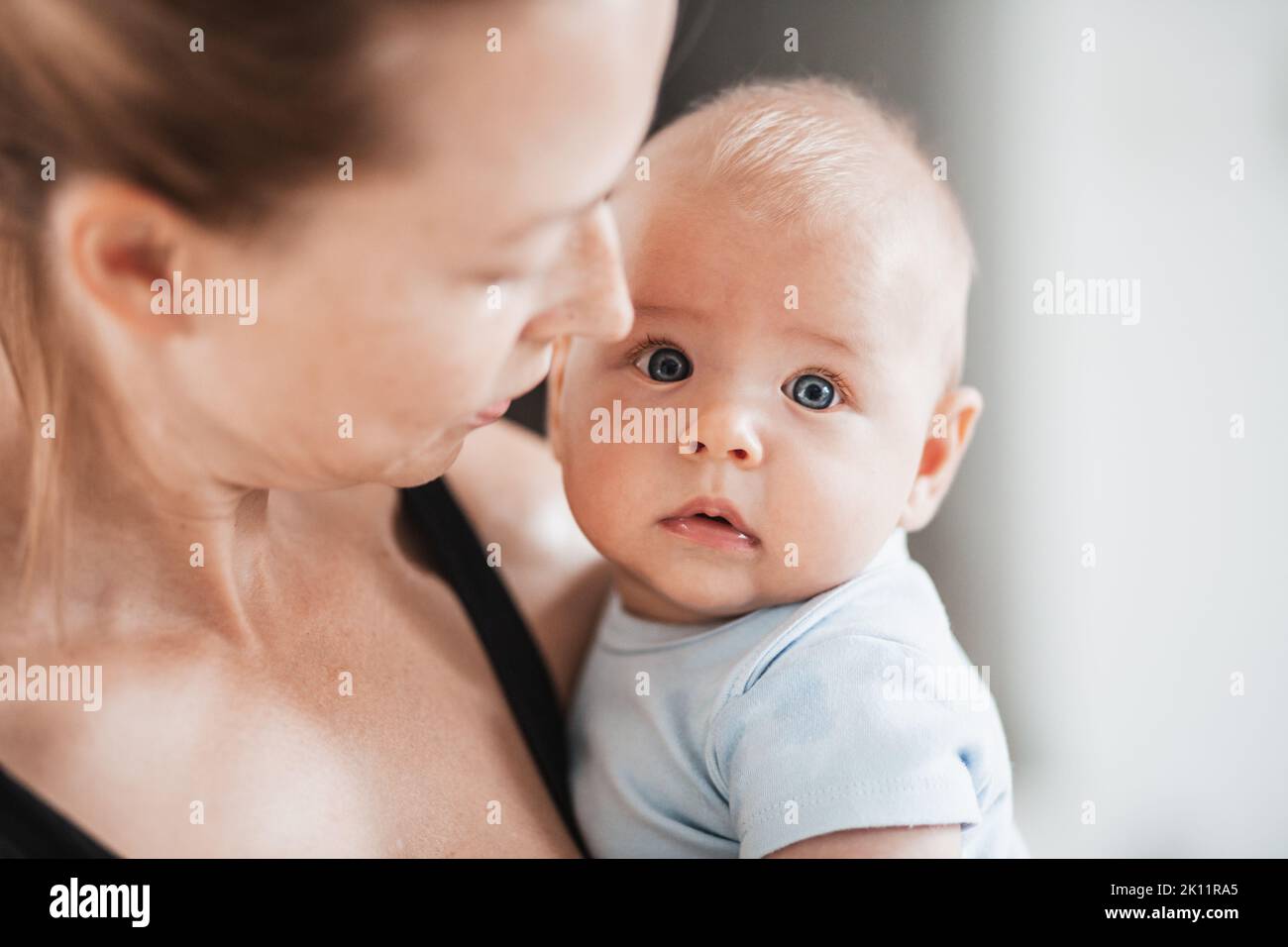 Portrait of sweet baby resting in mothers arms, looking at camera. New mom holding and cuddling little kid, embracing child with tenderness, love Stock Photo