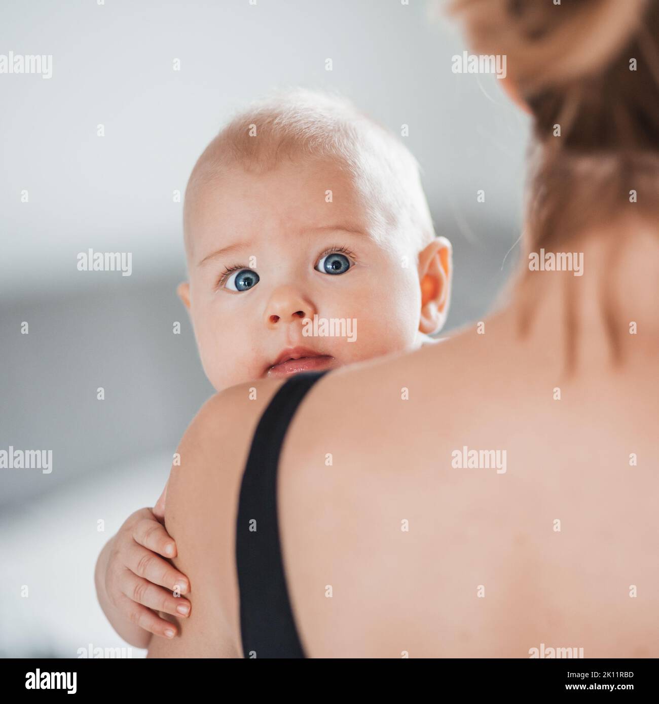 Portrait of sweet baby resting in mothers arms, looking at camera, touching mama shoulder. New mom holding little kid, embracing child with tenderness Stock Photo