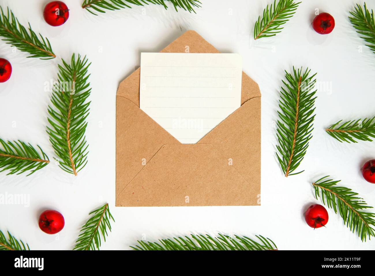 Close-up craft envelope, sheet of paper, letter to Santa. Frame of fir or spruce branch and red berries on white background. Christmas greeting card Stock Photo
