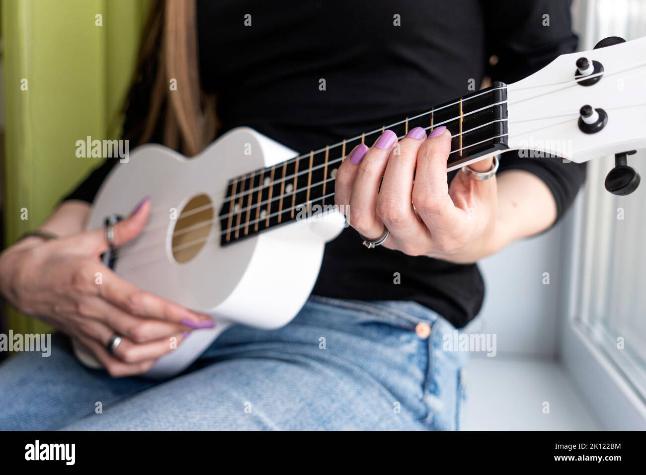 Girl sitting at home learns to play ukulele using online lessons. Stock Photo
