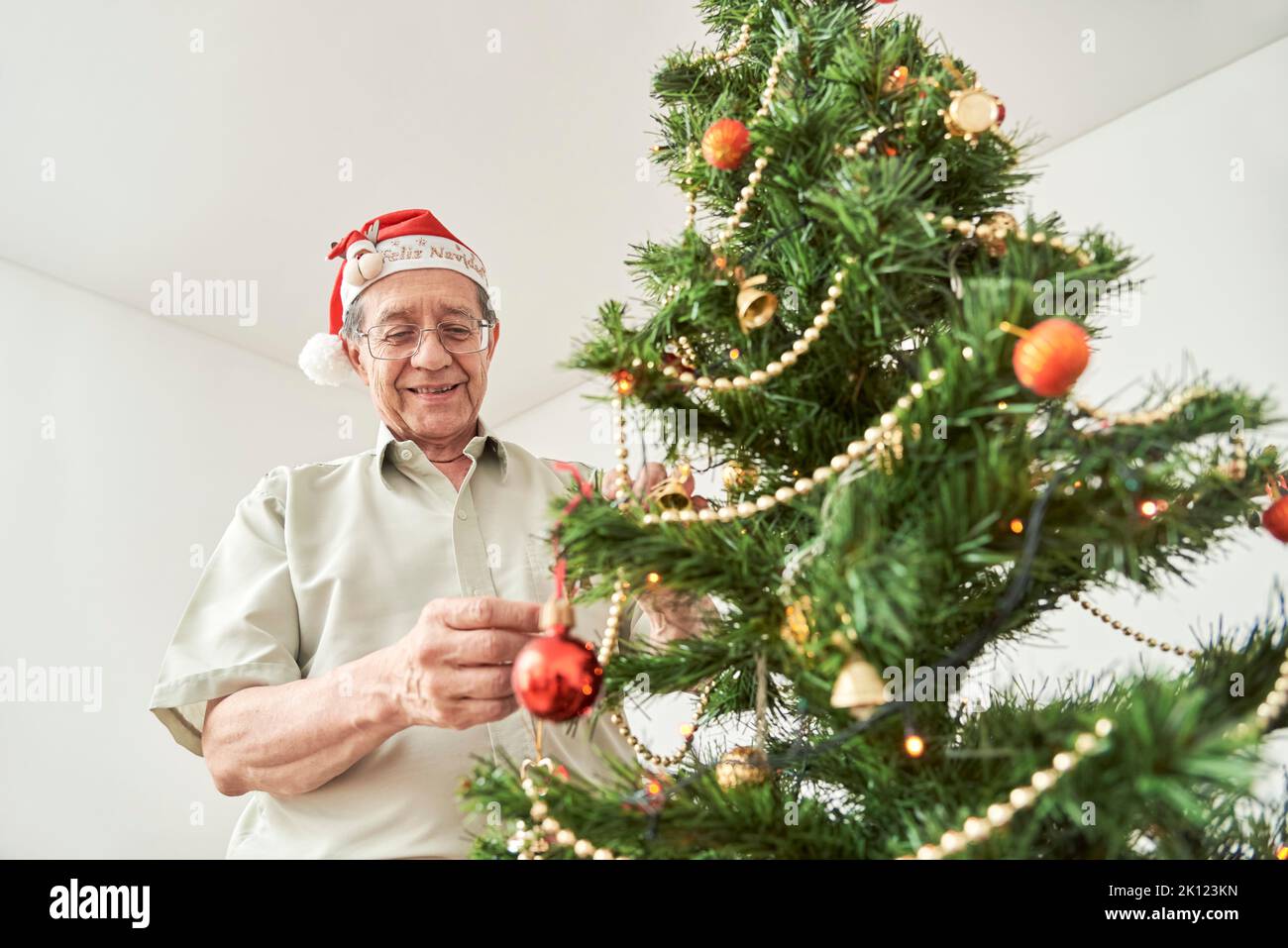 Happy senior hispanic man smiling while decorating a Christmas tree at home wearing a red Santa Claus hat with the text Merry Christmas. The happiness Stock Photo