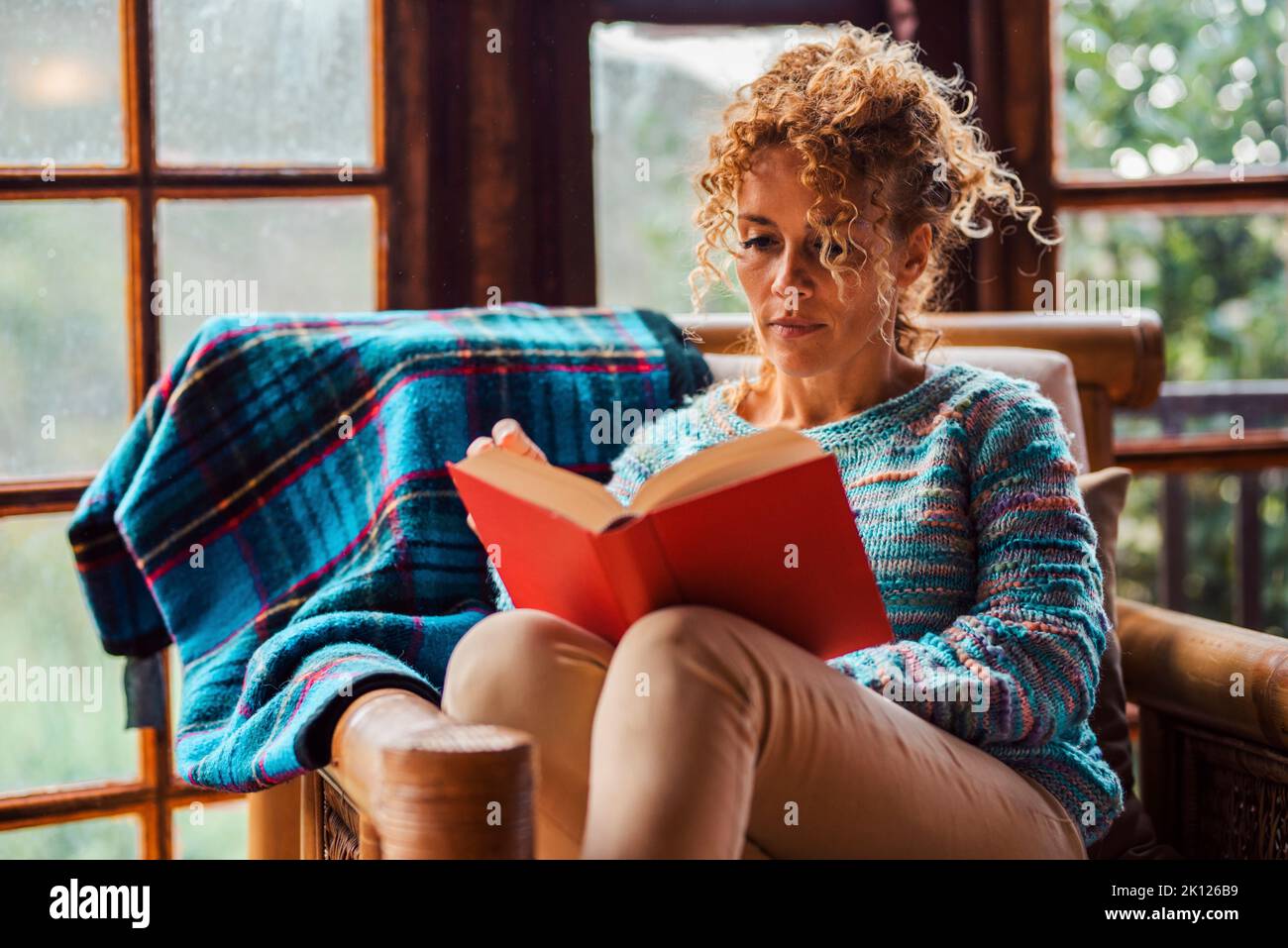 One serene woman at home relaxed sitting on a chair reading a book. Education and indoor leisure activity at home for happy single female people. Dail Stock Photo