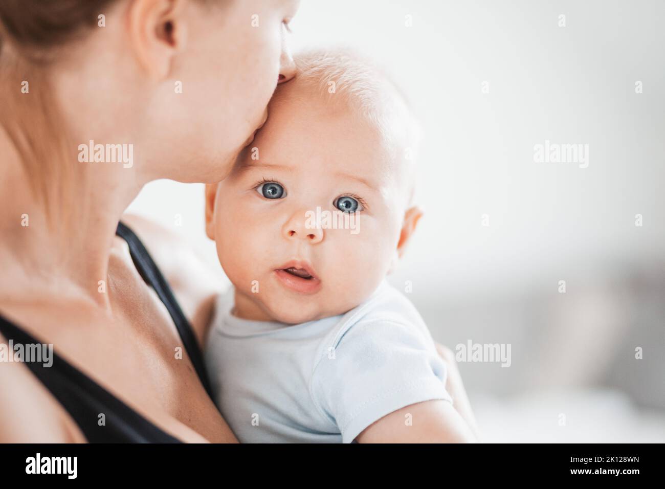 Portrait of sweet baby resting in mothers arms, looking at camera. New mom holding and kissing little kid, embracing child with tenderness, love, care Stock Photo