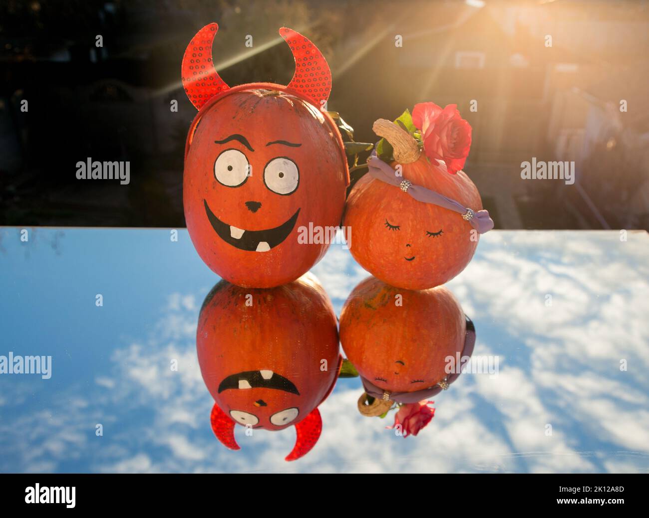 two beautiful orange pumpkins with a painted Halloween face stand on a mirror reflecting the blue sky, backlit by setting sun. preparing for Halloween Stock Photo
