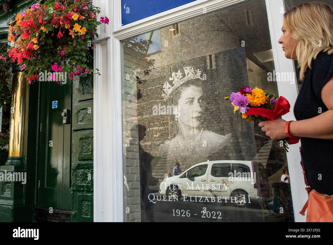 Windsor, UK. 14th September, 2022. A woman carrying flowers passes a tribute to Queen Elizabeth II displayed in a shop window. Queen Elizabeth II, the UK's longest-serving monarch, died at Balmoral aged 96 on 8th September after a reign lasting 70 years and will be buried in the King George VI memorial chapel in Windsor following a state funeral in Westminster Abbey on 19th September. Credit: Mark Kerrison/Alamy Live News Stock Photo