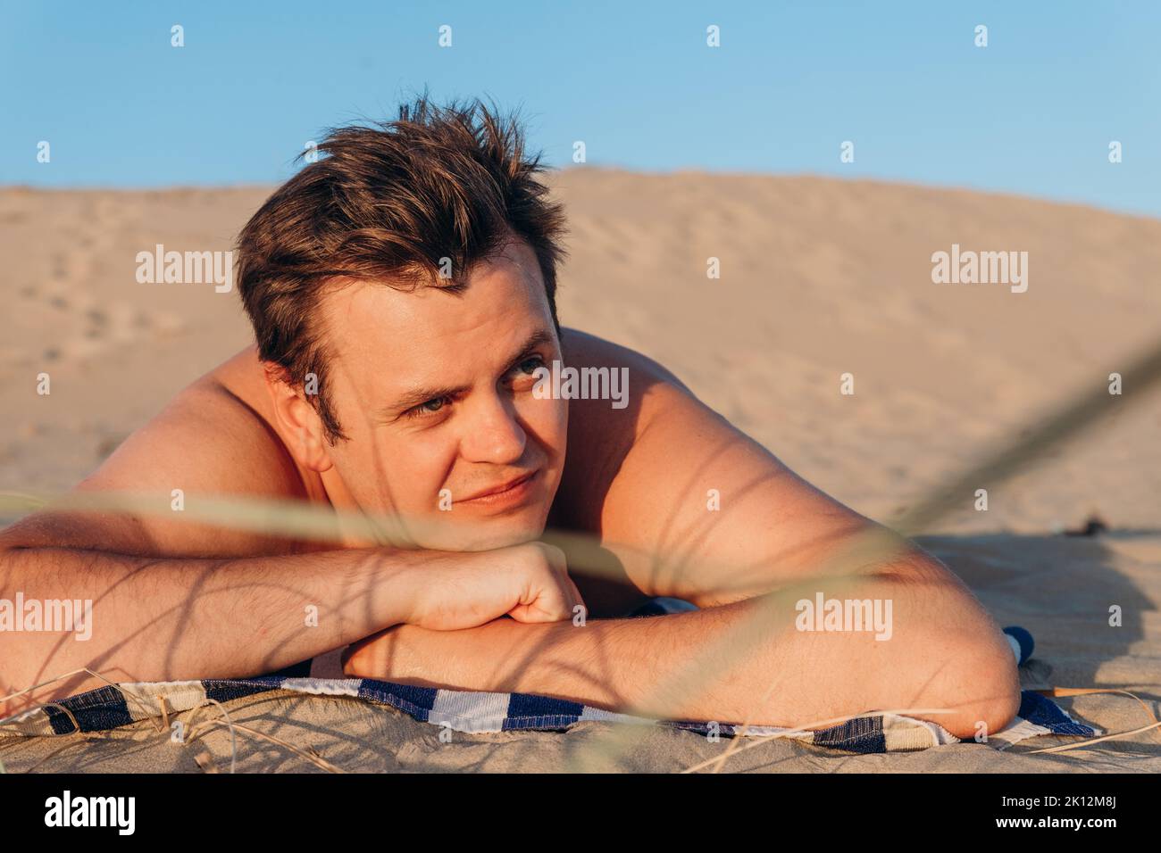 Looking away young 32 years old Caucasian man lies on striped towel and sunbathes on sandy beach on sand dune. Close-up summer headshot lifestyle thre Stock Photo