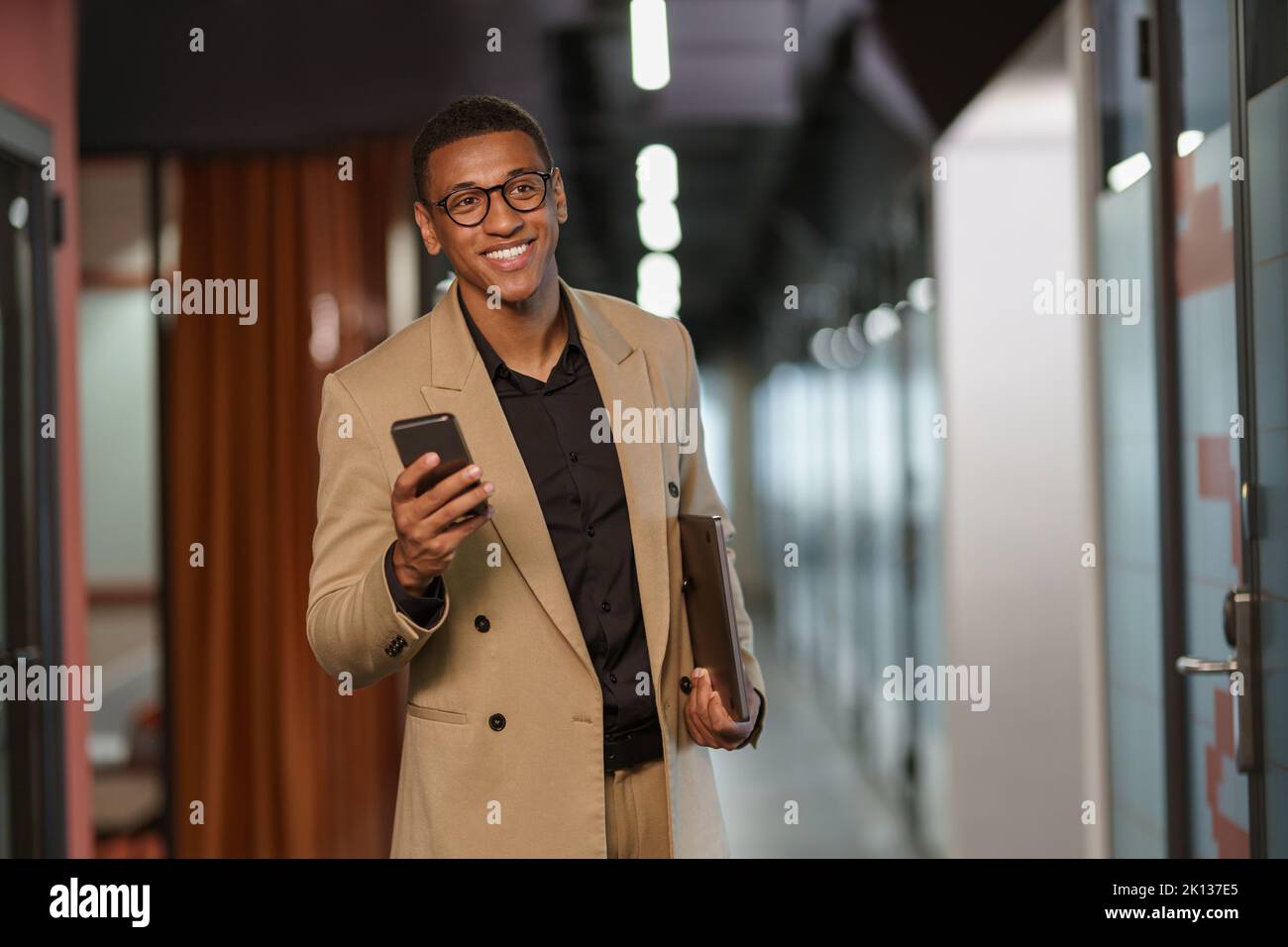 Handsome smiling african businessman using phone while standing in modern coworking Stock Photo