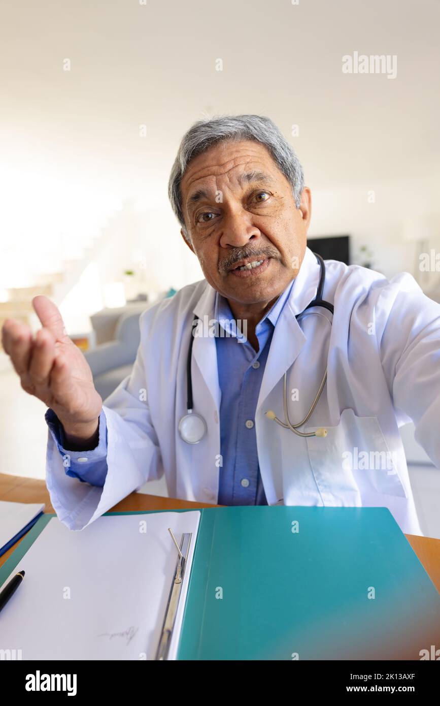 Verical image of senior biracial male doctor sitting at desk talking during video call consultation Stock Photo