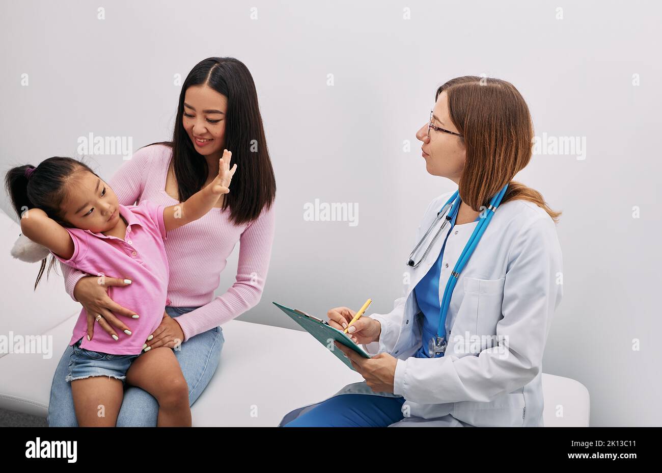 Beautiful Korean girl sitting next to her mother at pediatrician's appointment and showing doctor how old she is. Consultation, kid health Stock Photo
