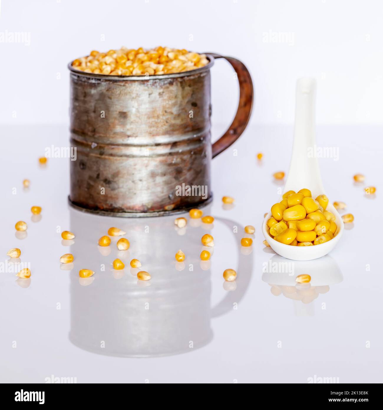 Cooked corn kernels on a white ceramic spoon with a metal container full of raw corn kernels in the background Stock Photo