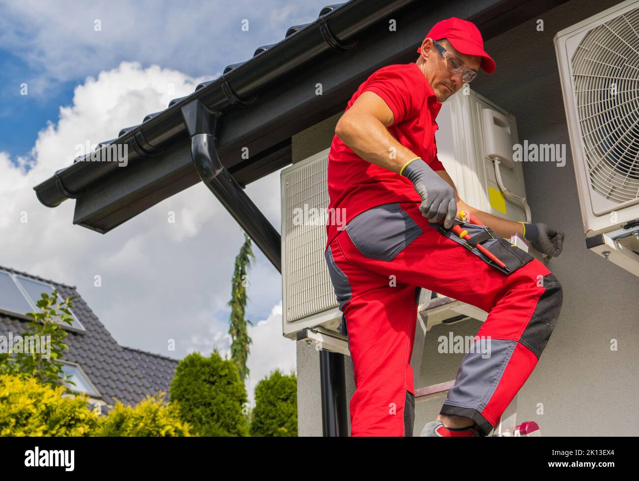 Modern Home Heating Technologies. Professional Caucasian HVAC Worker in His 40s Maintain Heat Pump Repair From a Ladder. Stock Photo
