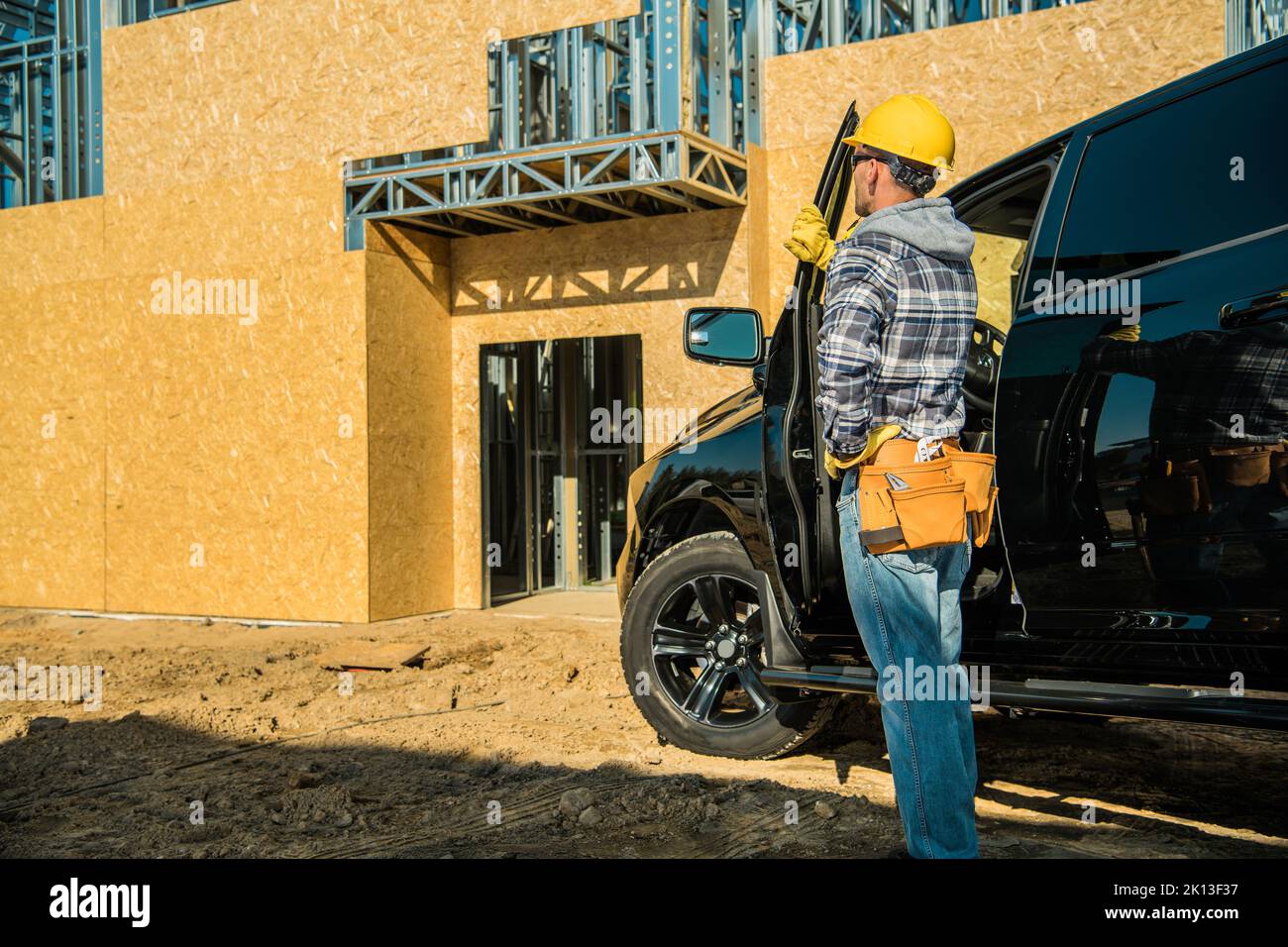 Caucasian Contractor Wearing Yellow Hard Hat and Tool Belt Standing Next to Open Pickup Truck Door Looking Ahead at Big Residential Building Under Con Stock Photo
