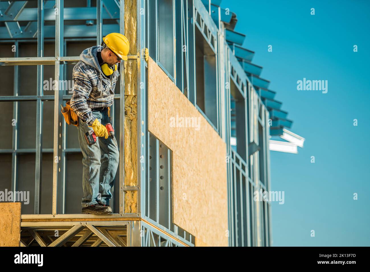 Construction Worker with Electric Cordless Screwdriver in His Hand Fearlessly Standing on the Non-Secured Wall of the House Steel Frame Installing Woo Stock Photo
