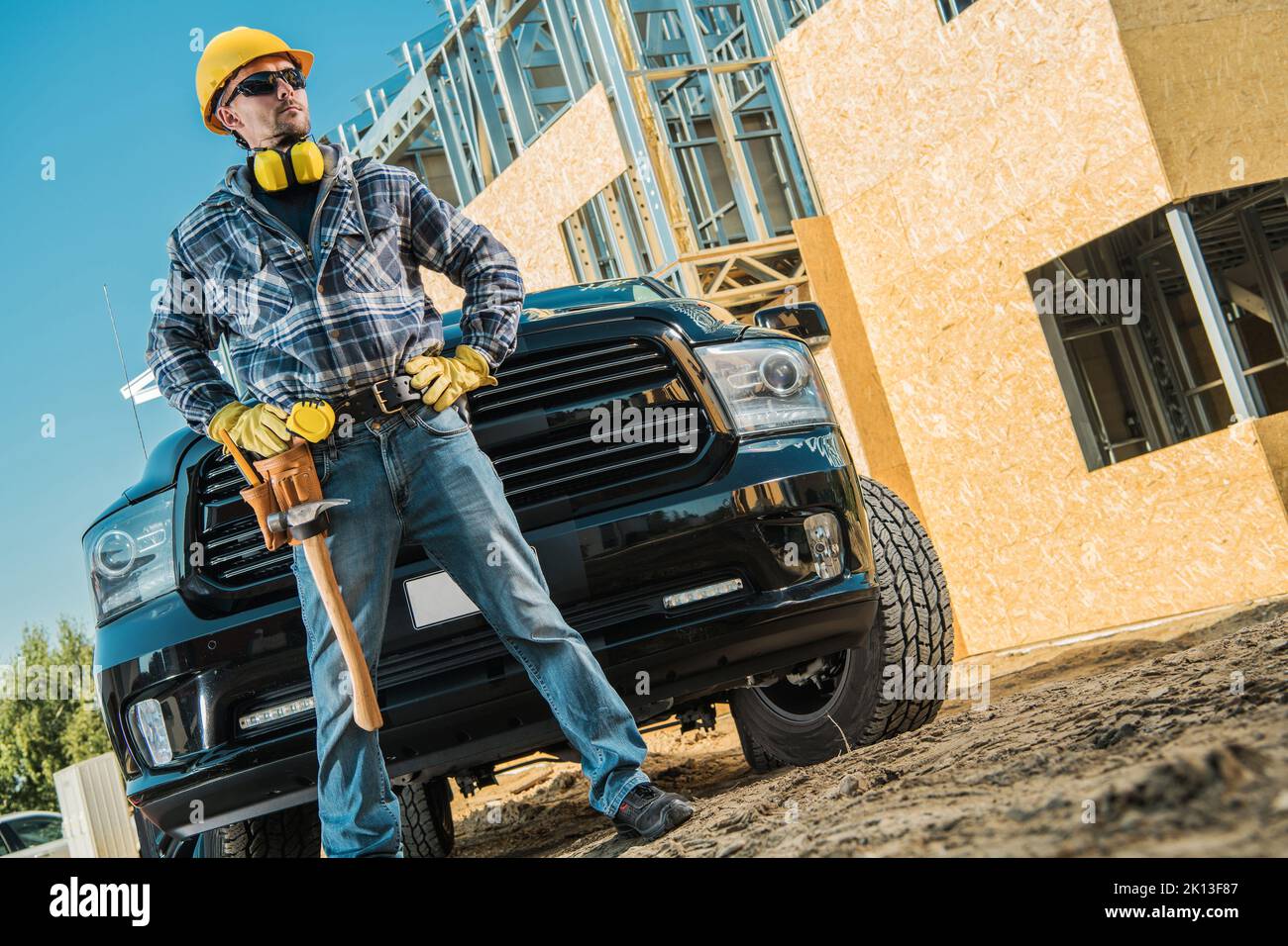Caucasian Middle Aged Builder Proudly Standing in Front of Black Pickup Truck. Equipped with Hard Hat, Safety Headphones and a Hammer Hanging From His Stock Photo