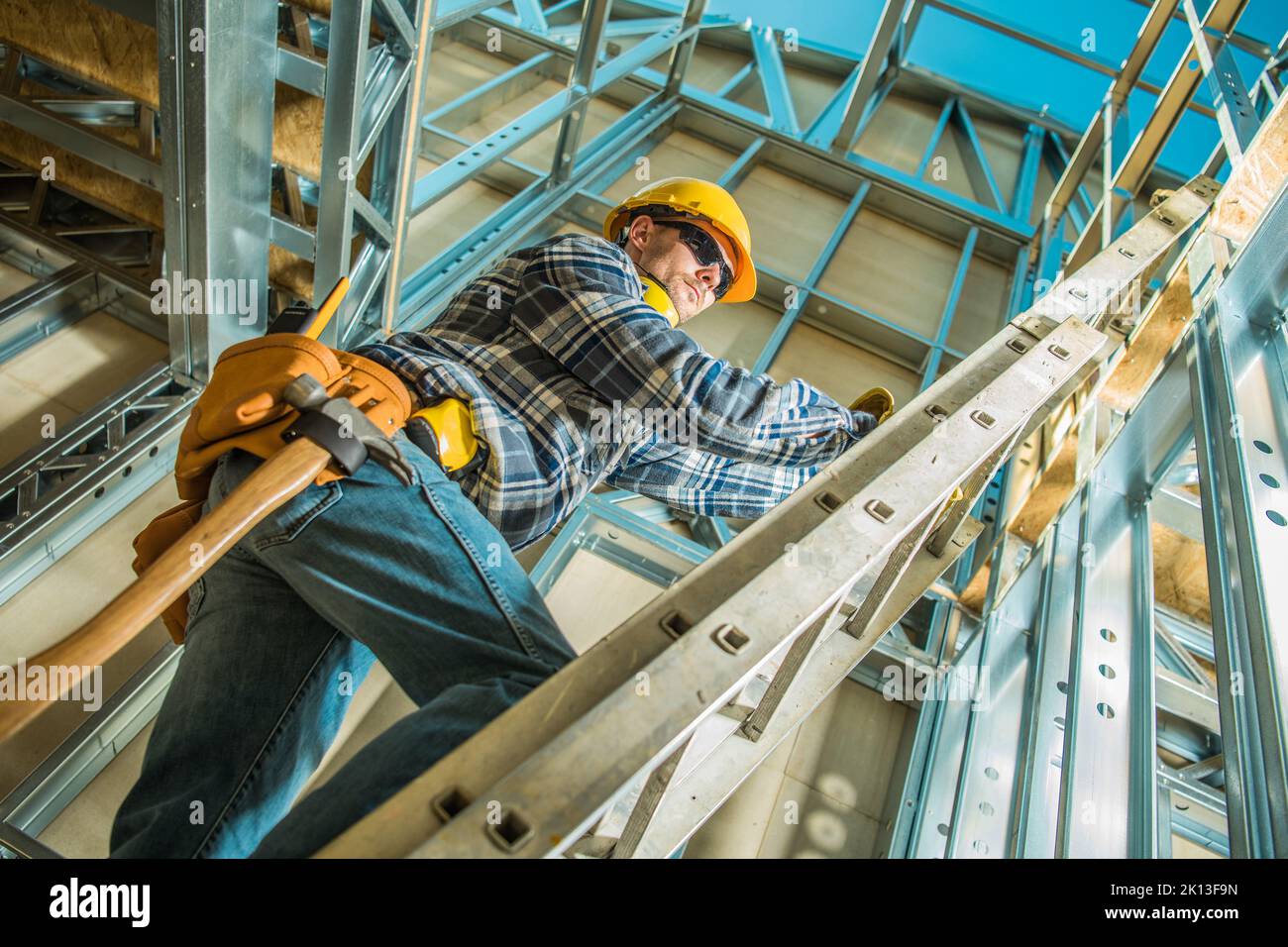 Construction Worker with a Hammer Hanging on His Tool Belt Stepping Up the Extension Ladder During Working on Building Steel Skeleton Frame of the Hou Stock Photo