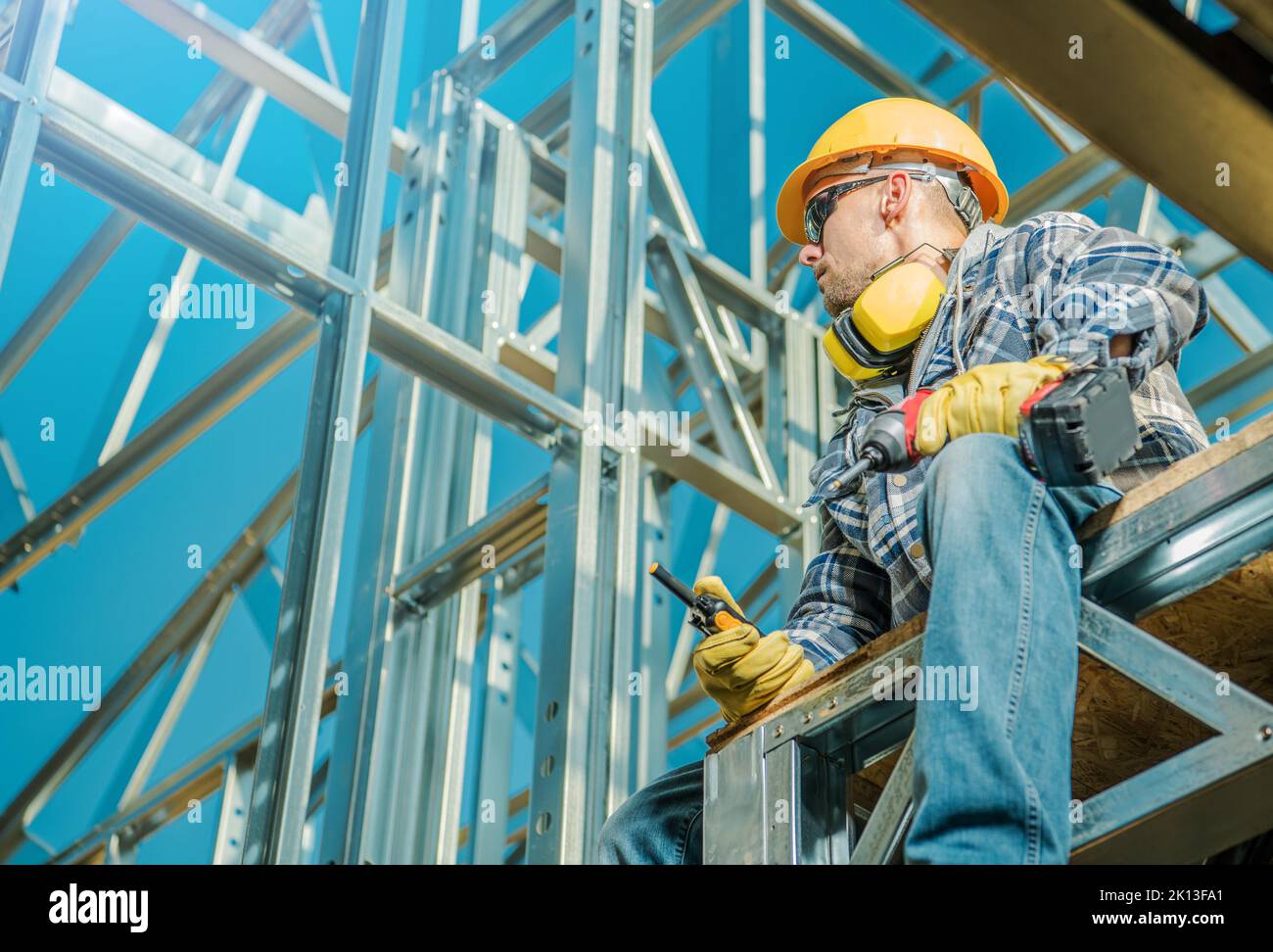 Caucasian Contractor with Walkie-Talkie in One Hand and Electric Screwdriver in the Other Sitting on the Construction Site. Steel House Skeleton Frame Stock Photo
