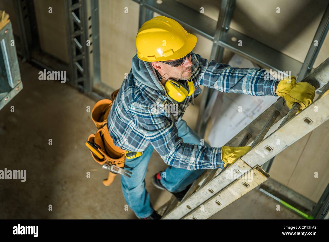 Top View Photo of Professional Contractor in Yellow Hard Hat Climbing the Ladder at the Construction Site. Building Tools and Equipment in Use. Stock Photo