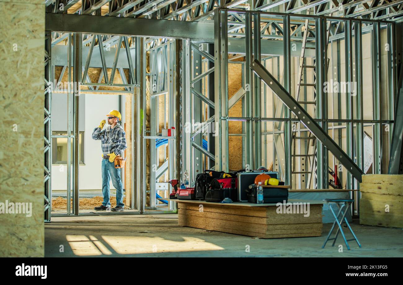Caucasian Construction Worker Standing on Building Site Surrounded by Steel House Frame with Wooden Elements. Wearing Yellow Safety Hard Hat and Commu Stock Photo
