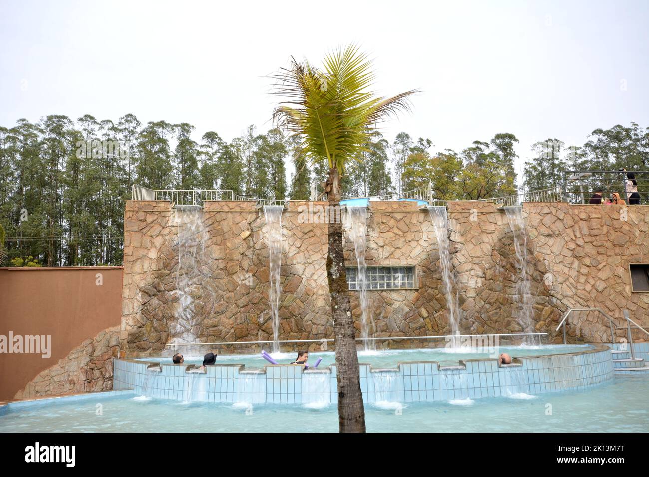 Tourists in a small modern spa pool with hot water stream in a luxury resort with waterfall or rock cascade on a day with a blue cloudy sky. Wide-ang Stock Photo