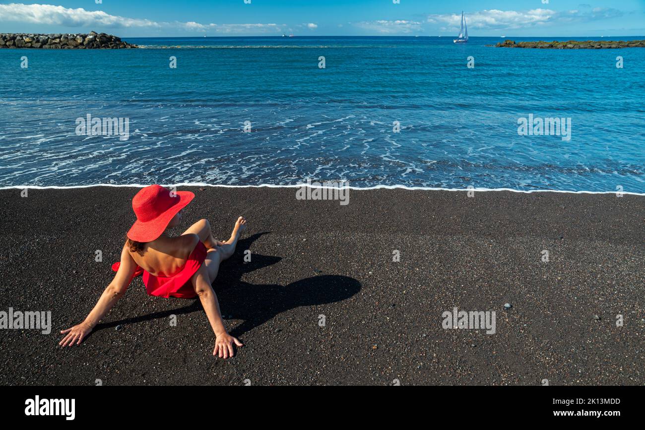 Woman in red swimming suit on the black ocean sandy beach. Calm ocean water and pure blue sky at the background. Stock Photo