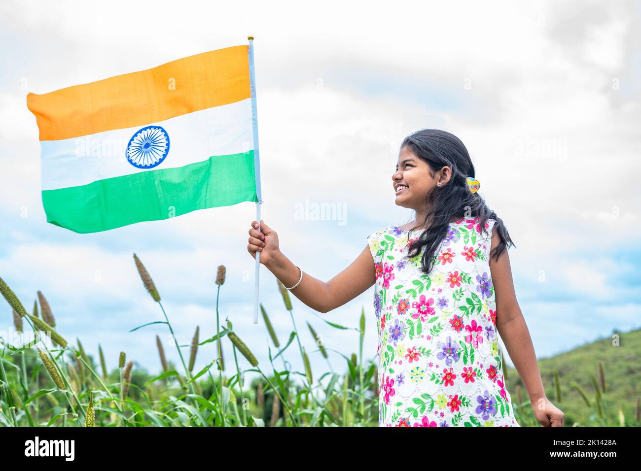 happy smiling girl kid with indian flag in hand running at pearl millet farm land near green mountain - concept of independence, liberty and Stock Photo