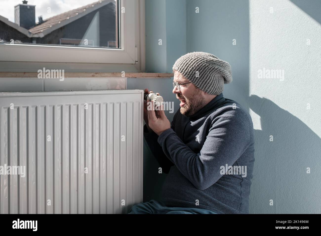 Irritated man in warm clothes turns the radiator thermostat to minimum. Concept of crisis and increase in fuel costs. Preparing for a cold winter. Stock Photo