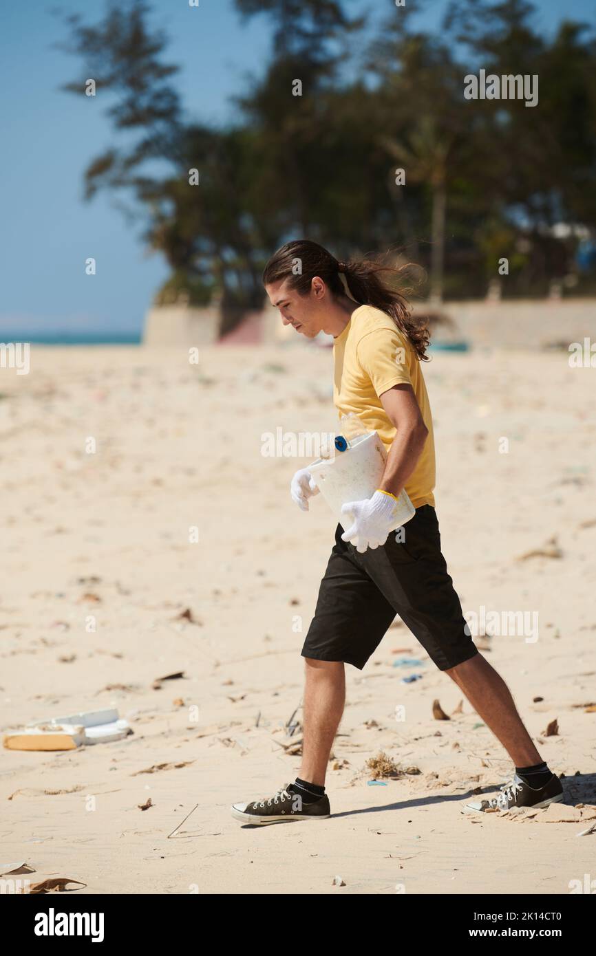Young man with plastic bucket walking on beach and collecting garbage