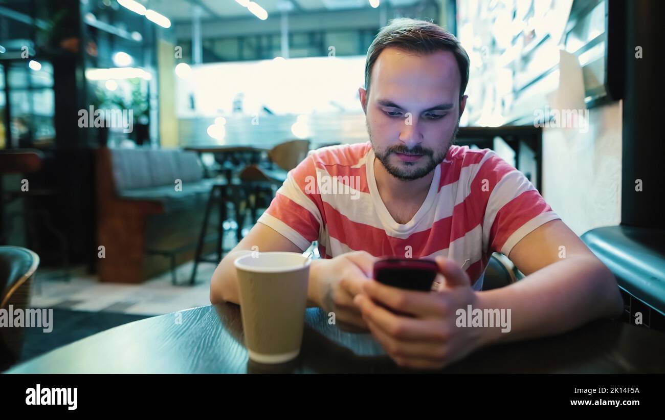 Young attractive man with a beard uses a smartphone in a cafe. Stock Photo
