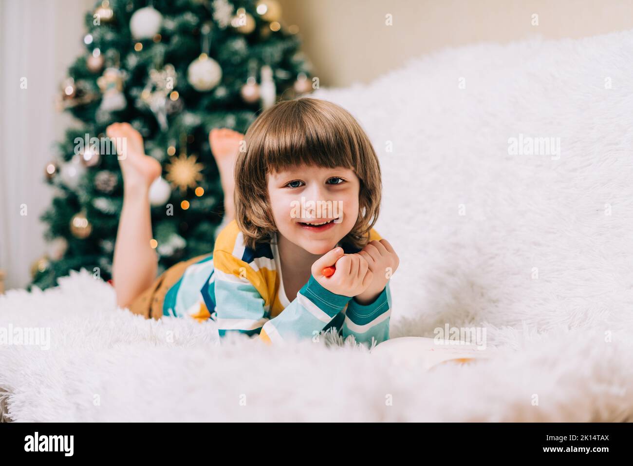 Smiling Kid boy lying on couch and writing the letter to dear Santa at home near Christmas tree. Child wish list. Dreams of a Christmas gifts. Merry Stock Photo