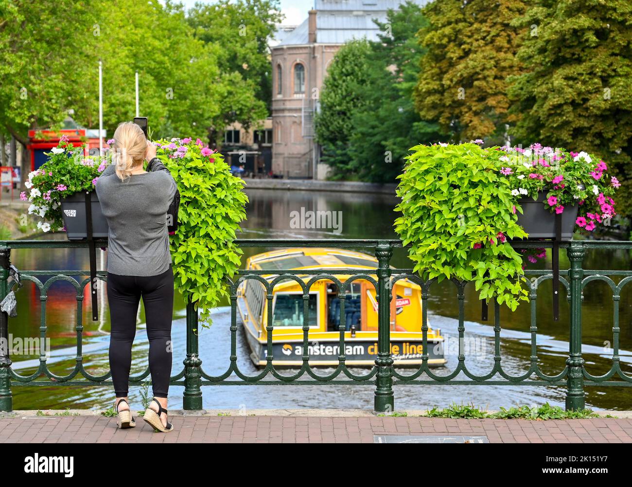 Amsterdam, Netherlands - August 2022: Person on a bridge with flower baskets taking a picture of a boat on one of the city's canals Stock Photo