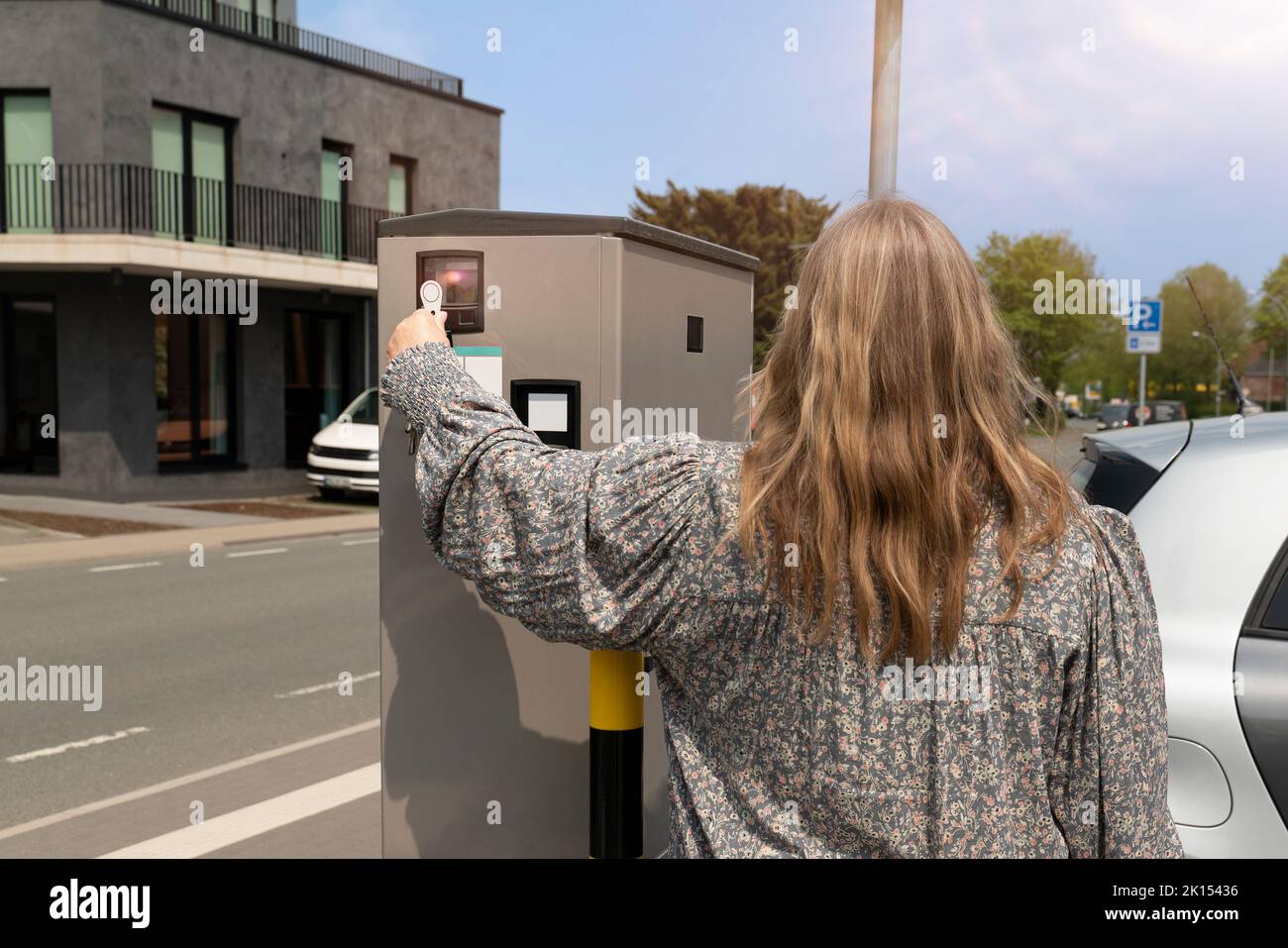 A woman is standing at an e car charging station. She is making a contactless payment. In the background is a street with traffic. Stock Photo