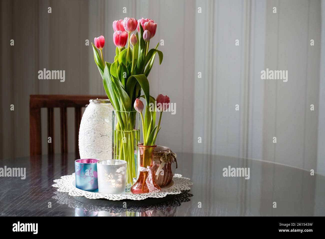 Long stemmed pink tulips with several glass jars on a round wooden table. In the background is a chair and striped wallpaper. Antique romantic look. Stock Photo