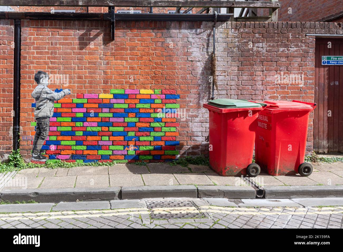 Street art mural by Hendog, an anonymous artist, depicting a young boy building a colourful lego wall, in Winchester, Hampshire, UK, next to bins. Stock Photo