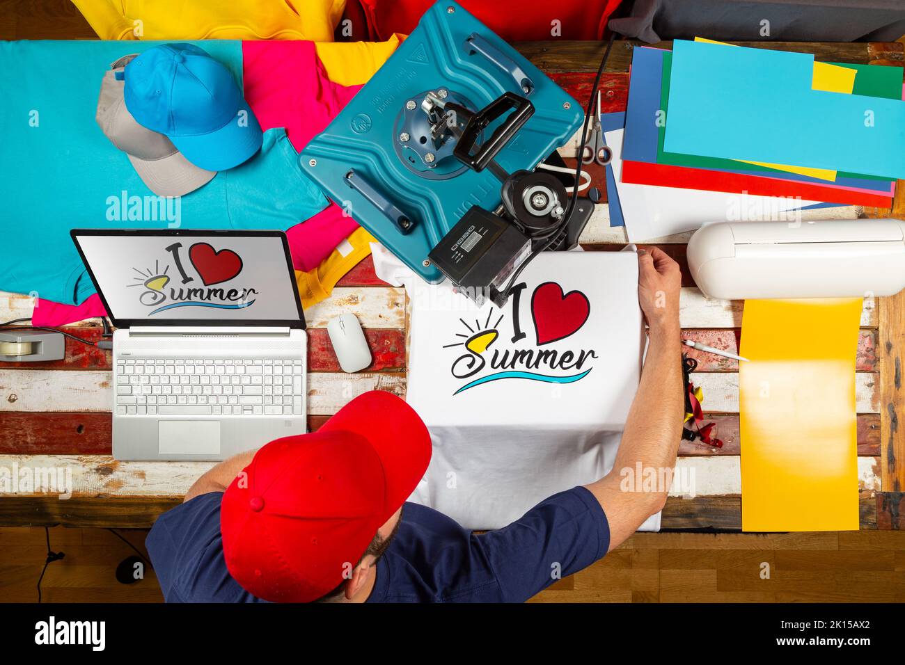 Young man working on heat transfer shirt press to manufacture custom DIY design shirts with notebook computer and cutting machine. Startup fashion des Stock Photo