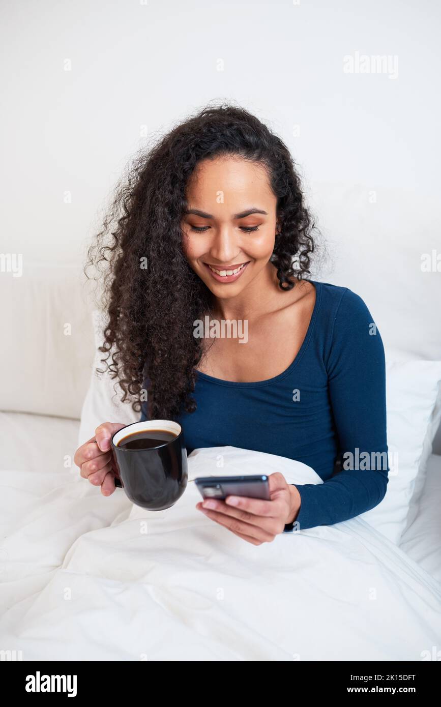 A young multiethnic woman drinks coffee in bed while on her phone