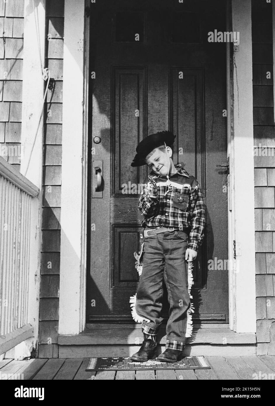Six year old, Lewis E. Raymond, III, dressed as a cowboy brandishes his toy gun on the porch of his home. Many children participate in a form of role-playing known as make believe, wherein they adopt certain roles such as doctor and act out those roles in character. Sometimes make believe adopts an oppositional nature, resulting in games such as cops and robbers. Circa 1950-60s. Stock Photo